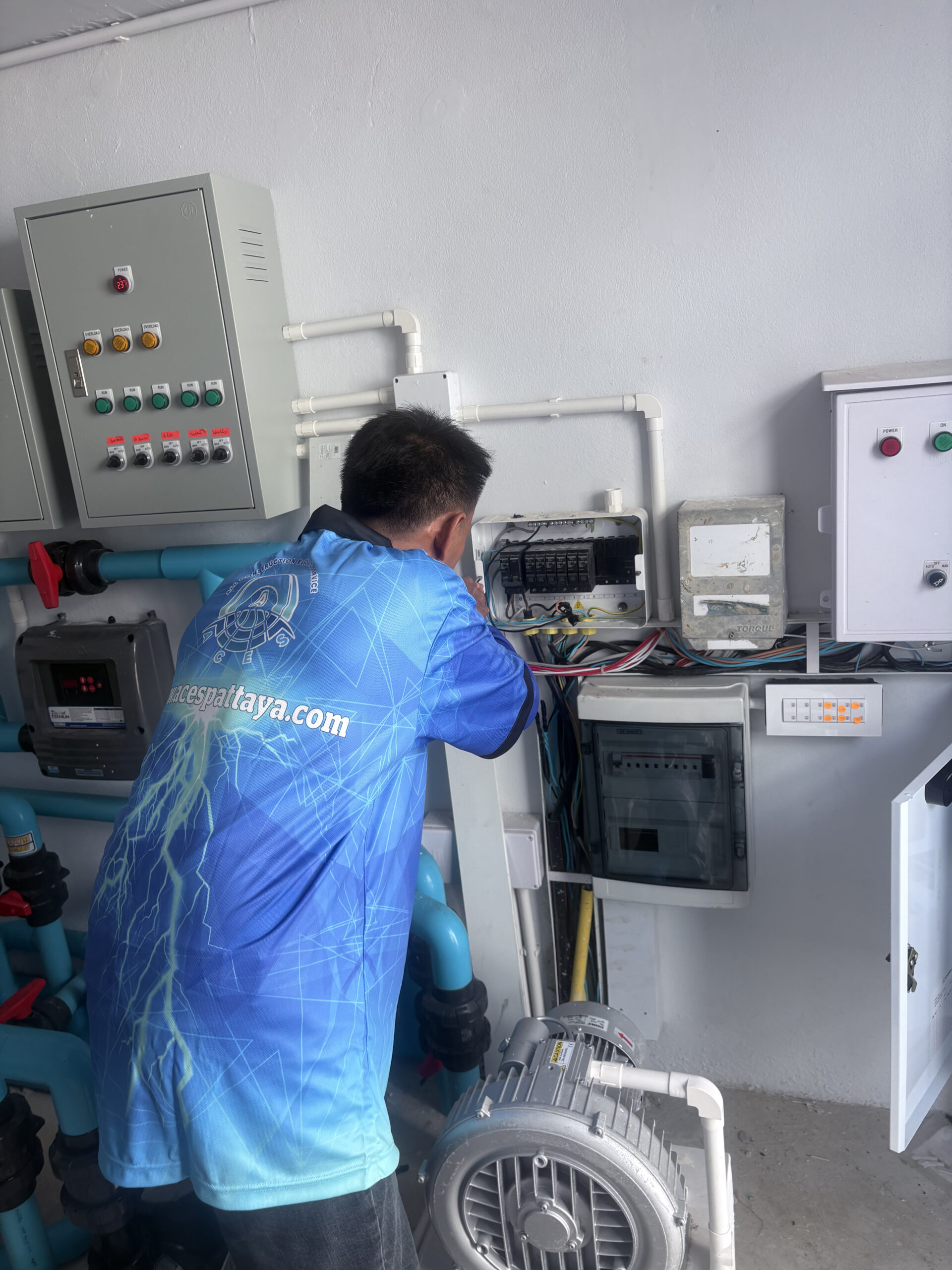 An ACES Pattaya technician wiring an electrical distribution board next to commercial pump control panels and industrial utility piping.