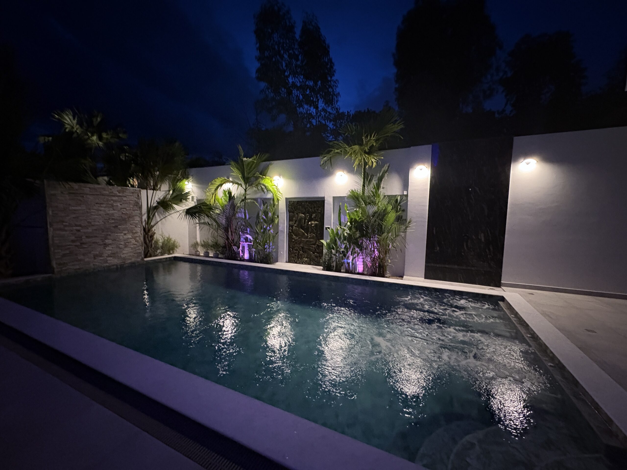 Luxury private swimming pool at night featuring custom outdoor wall lighting and colorful landscape uplighting in Pattaya.