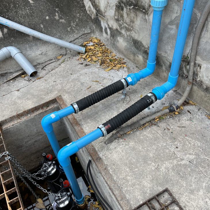 Top-down view into a concrete drainage pit featuring two heavy-duty industrial submersible sump pumps with lifting chains and blue PVC discharge piping in Pattaya.